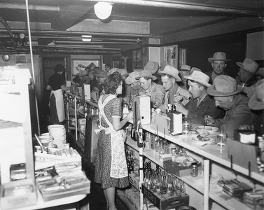 Two waitresses wearing dresses and floral aprons stand behind counter while men wearing cowboy hats eat at the bar. Behind the counter is a box of empty Nehi glass bottles. Possibly the Stockman's Cafe, or the restaurant in the basement of the Exchange Building. Thank you to Don Strack for generously sharing this photo, part of his extensive gallery.